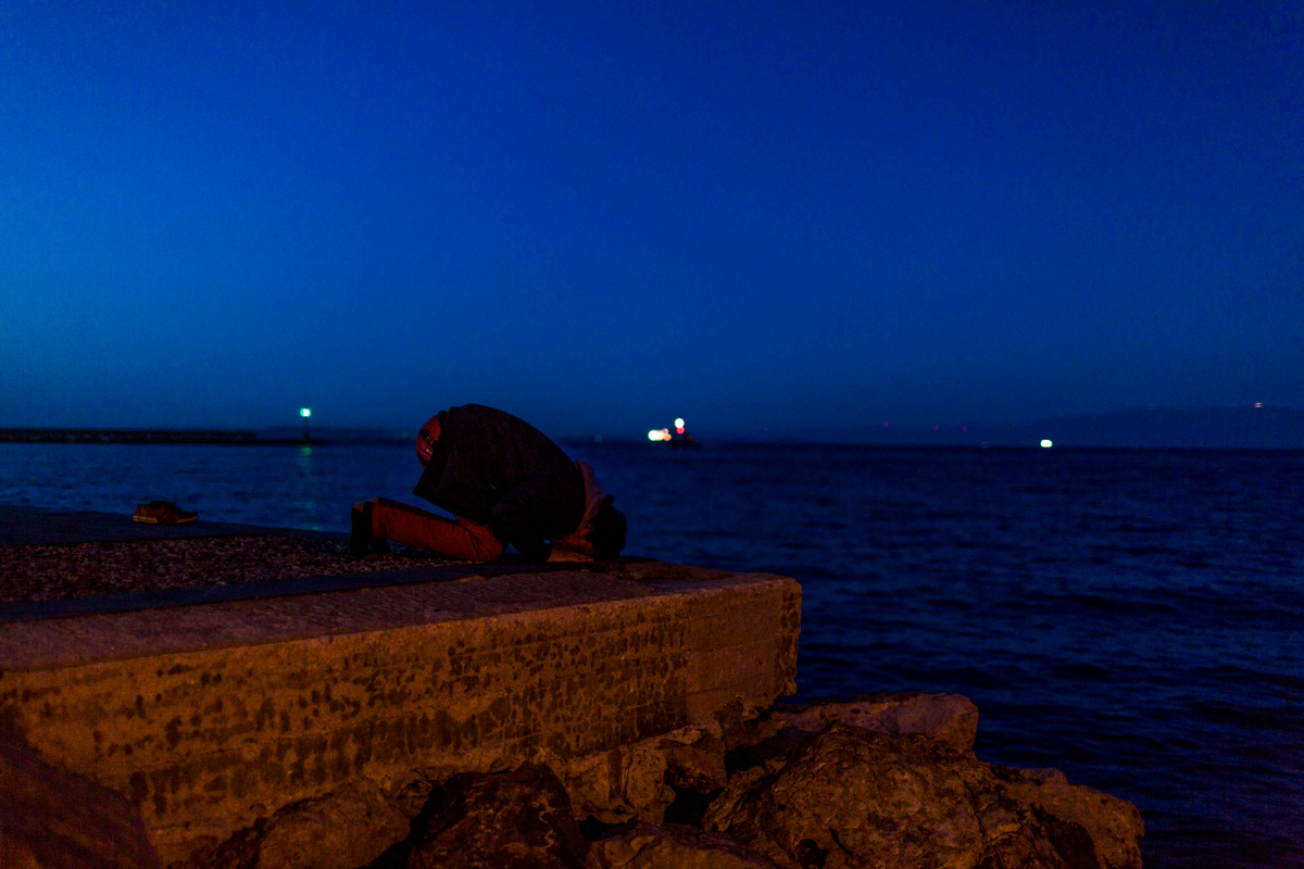 Port of Chios. Falah, a 26 year Pakistani refugee, pray on the harbour. In the background the Turkish coast.